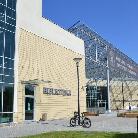 Fragment budynku Centrum Wykładowego PP na Piotrowie. Po lewej stronie widoczne wejście do obiektu, przy którym jest napis Biblioteka. Po prawej w głębi widoczne jest frontowe, główne wejście do budynku.