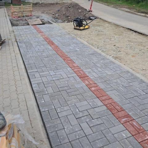 Chodnik przed tylnym wejściem do Centrum Sportu PP w trakcie przebudowy. Na pierwszym planie widoczna nowa kostka brukowa w kolorze szarym, przedzielona na środku, wzdłuż całej długości, wąskim pasem kostki w czerwonym kolorze. Z tyłu widać kopce materiałów budowlanych (piasek, żwir, kostka) ora urządzenie do ubijania powierzchni.