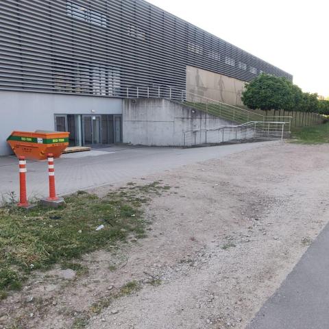 Zdjęcie przedstawia tylne wejście do budynku Centrum Sportu PP. Po prawej widać asfaltową drogę, do której od lewej przylega kawałek piszczystego terenu. Dalej po lewej widać utwardzoną powierzchnię przed drzwiami do budynku i sam budynek Centrum.