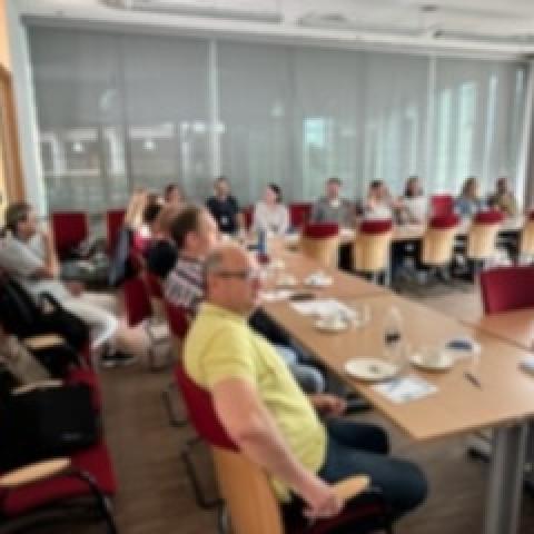 Sala wykładowa zapełniona uczestnikami szkolenia. Ludzie siedzą przy stołach ustawionych w ksztalt litery U. 