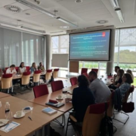 Sala wykładowa zapełniona uczestnikami szkolenia. Ludzie siedzą przy stołach ustawionych w ksztalt litery U. Na końcu sali, na tle okien, wisi ekran wyświetlający prezentację.