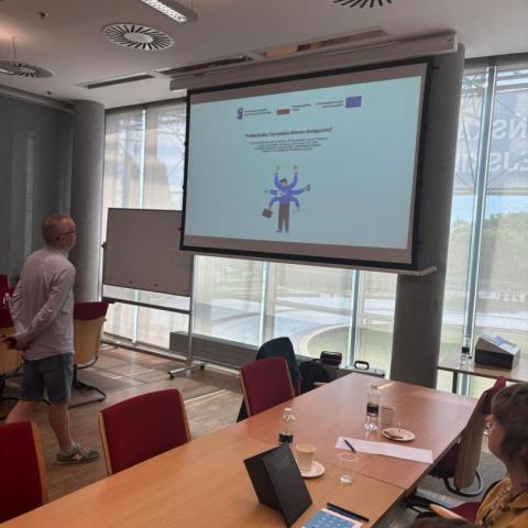 Sala konferencyjna. Stół, przy nim ludzie. W głębi ekran z prezentacją i prelegent