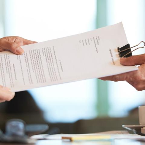 Grafika ozdobna. Na pierwszym planie widać dokument spięty klipsem przekazywany z rąk jednej osoby w ręce drugiej.