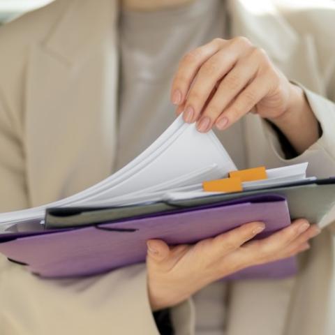 Grafika ozdobna. Kobieta trzyma w dłoni plik dokumentów. Drugą ręką przegląda plik kartek.