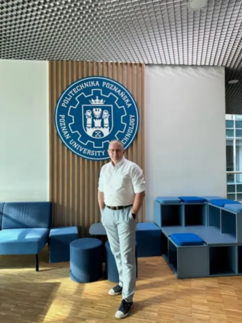Zdjęcie prowadzącego szkolenie Przemysła Staniszewskiego. Mężczyzna stoi na tle dużego logo Politechniki Poznańskiej, na korytarzu budynku tej uczelni.