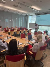 Sala konferencyjna. Stoły ustawione w kształcie litery C, przy nich ludzie. W głębi ekran z prezentacją i prelegent zwrócony przodem do słuchaczy